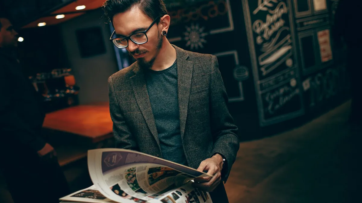 A man in a brown blazer reads a magazine in a dimly lit