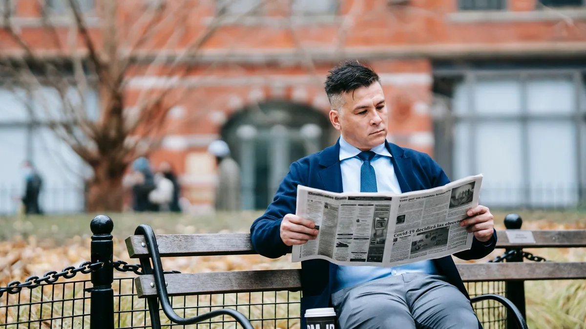 A man in formal attire sits on a park bench reading a ne
