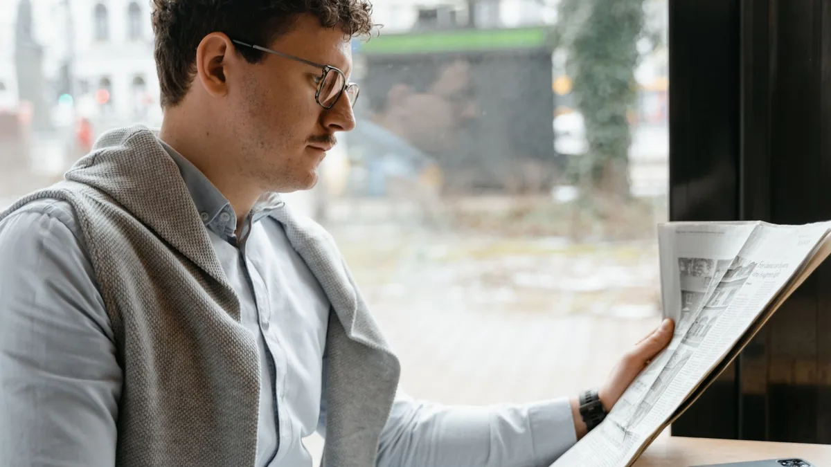 A man reading a newspaper by a window wearing glasses an