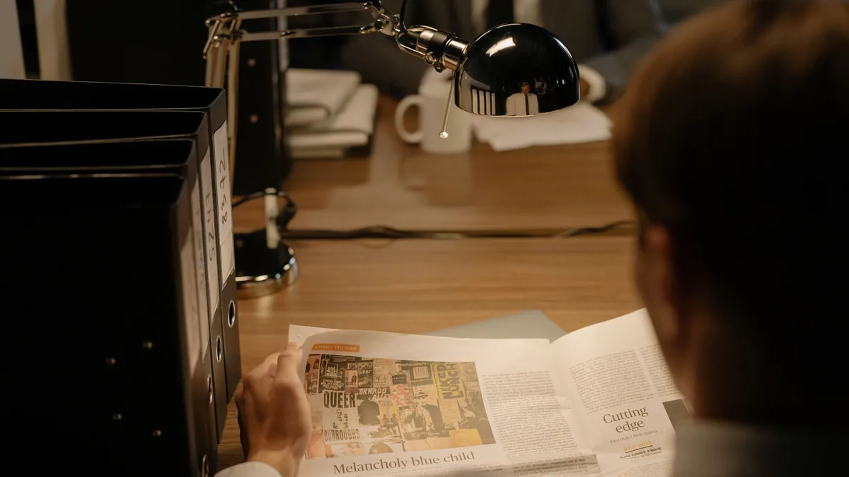 A person reads a newspaper at a desk with a lamp suggest