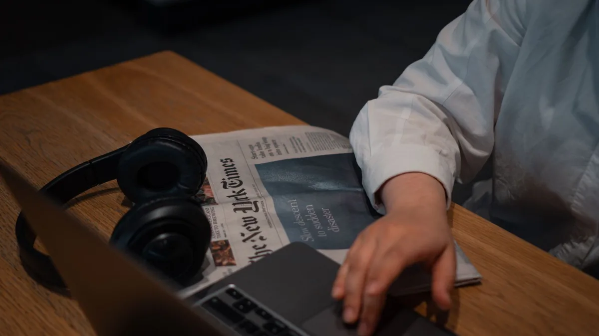 A person works on a laptop with headphones and a newspap