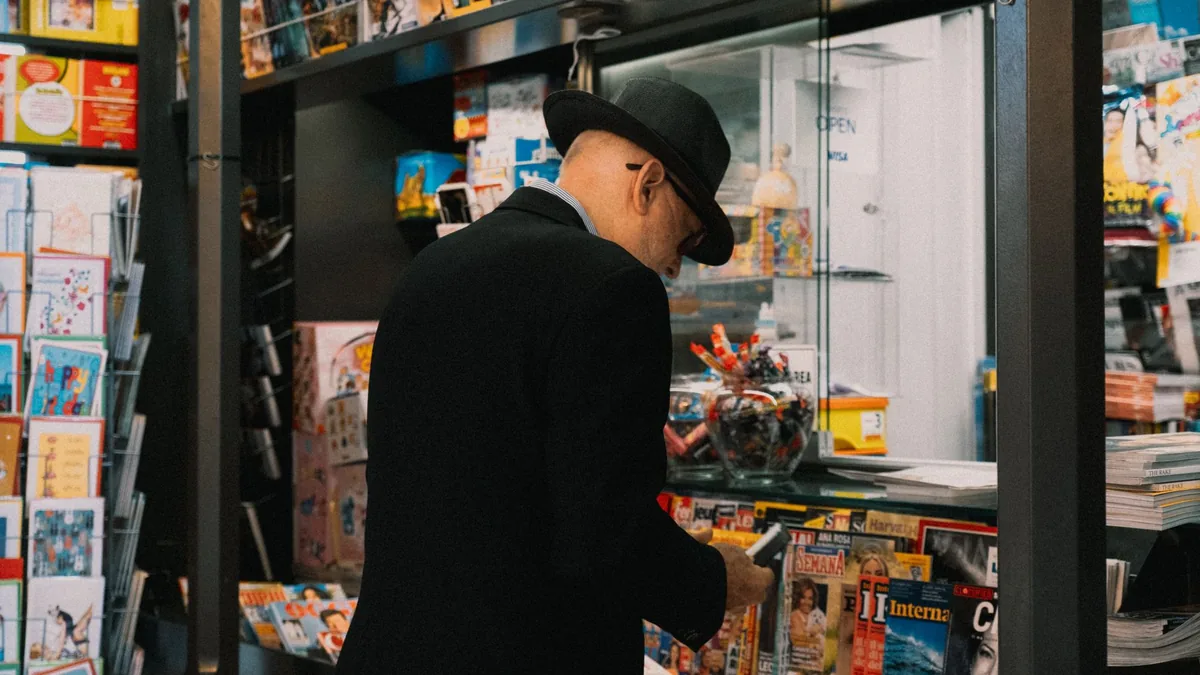 A senior man in a hat browsing magazines at a kiosk indo