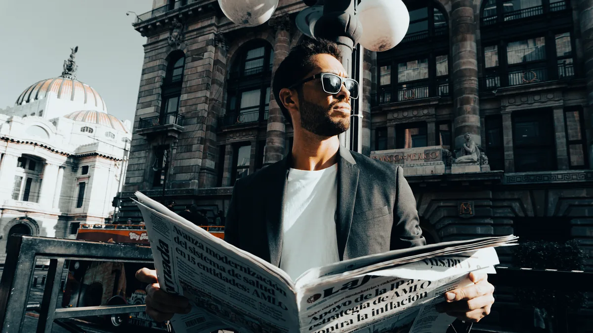 A stylish man in sunglasses reads a newspaper outside pa
