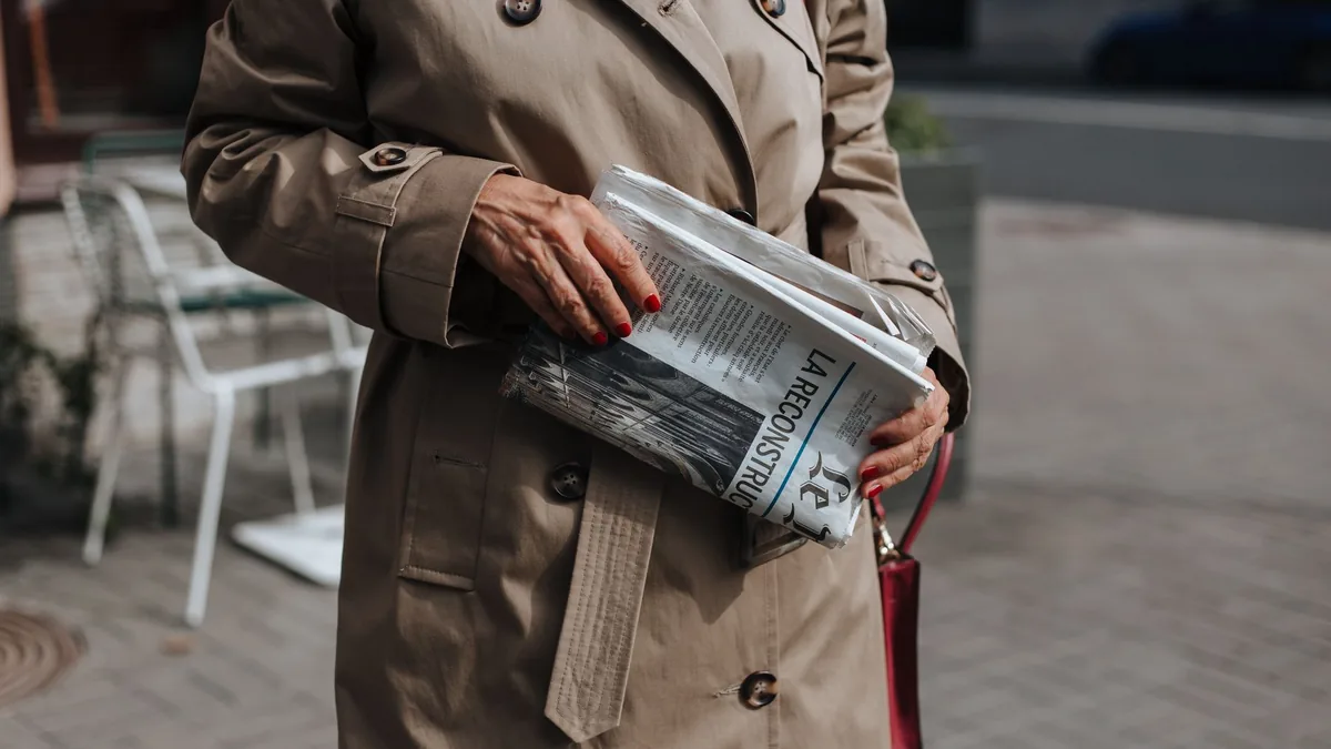 A woman in a trench coat holding a newspaper on a city s