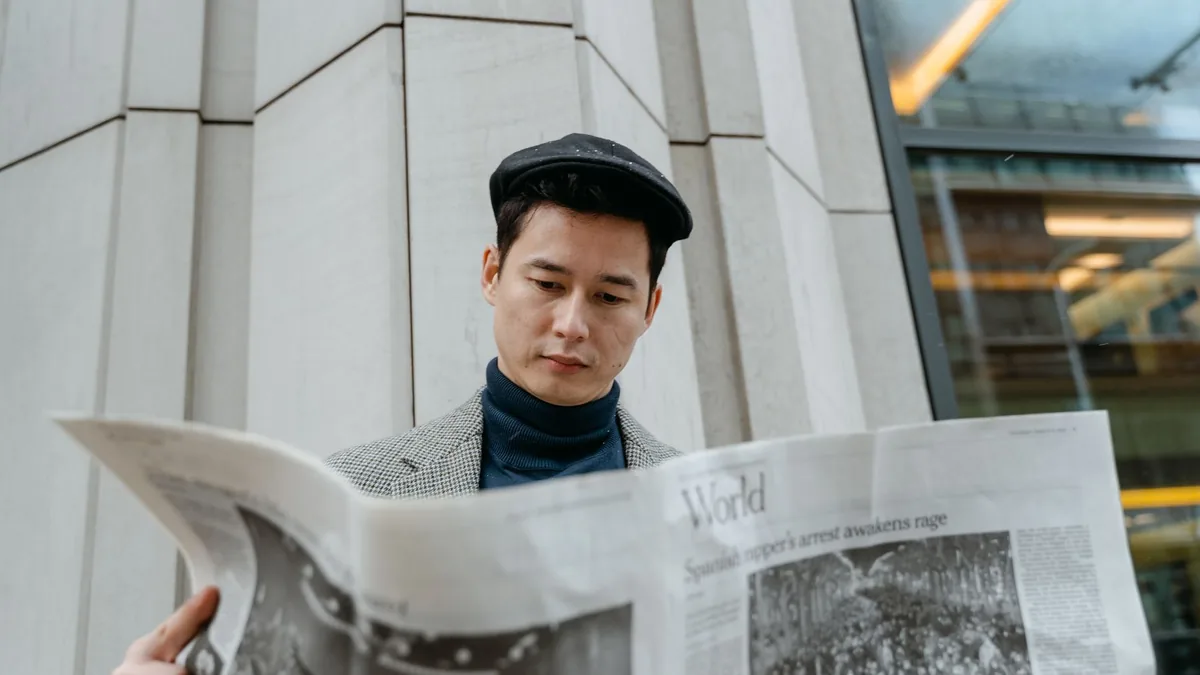 A young man in a flat cap and turtleneck reads a newspap