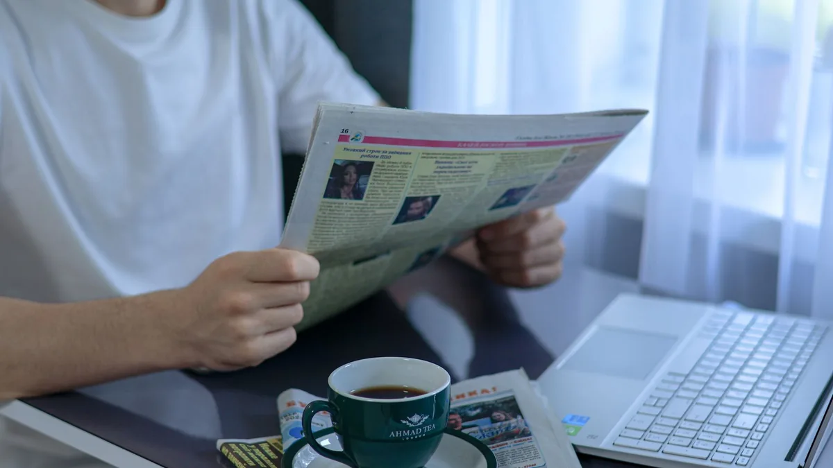 Adult enjoying coffee and newspaper at a desk with lapto