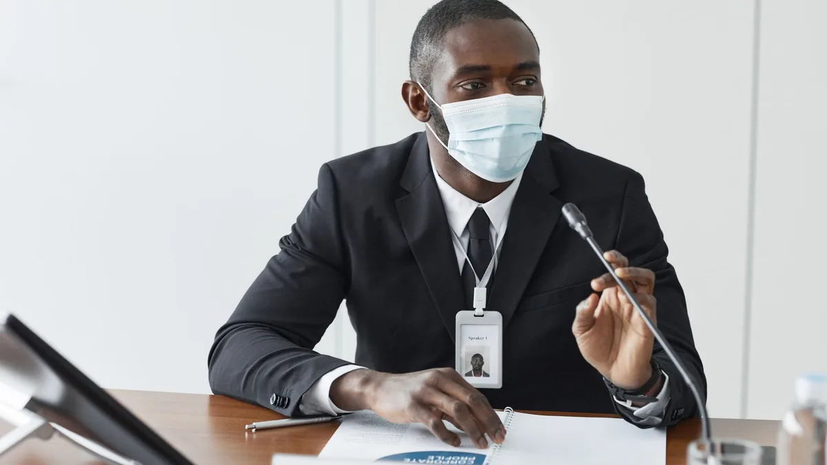 An adult male wearing a suit and face mask speaks at a c