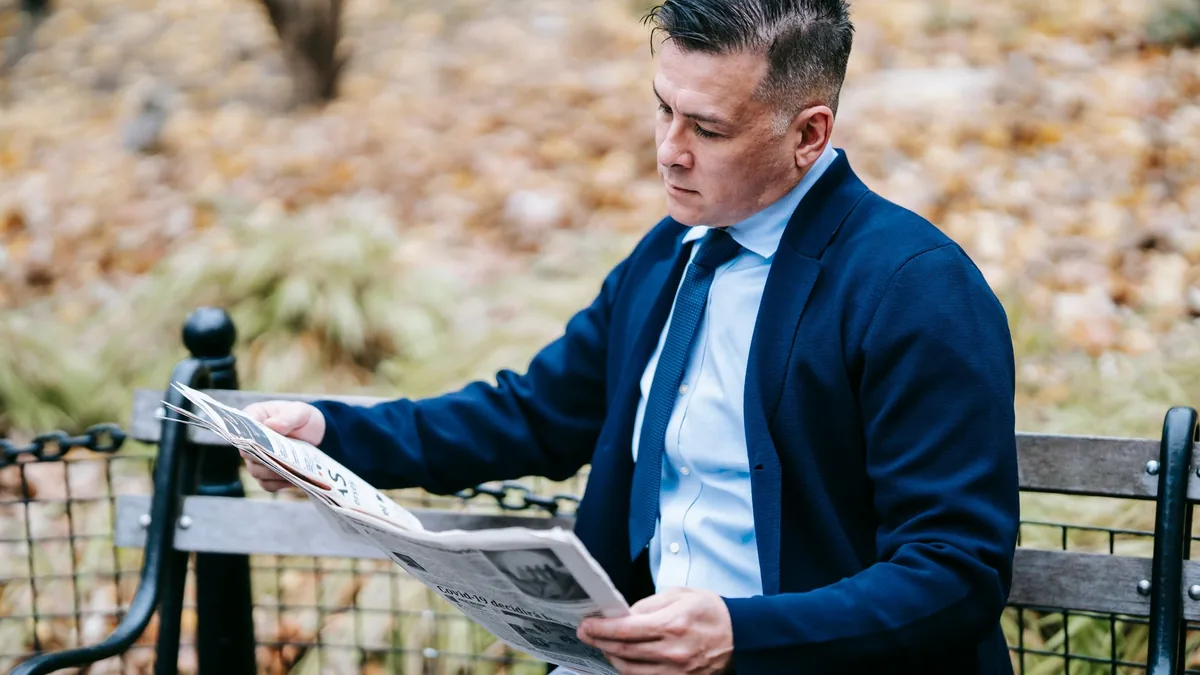 Businessman in blazer reads a newspaper on a park bench