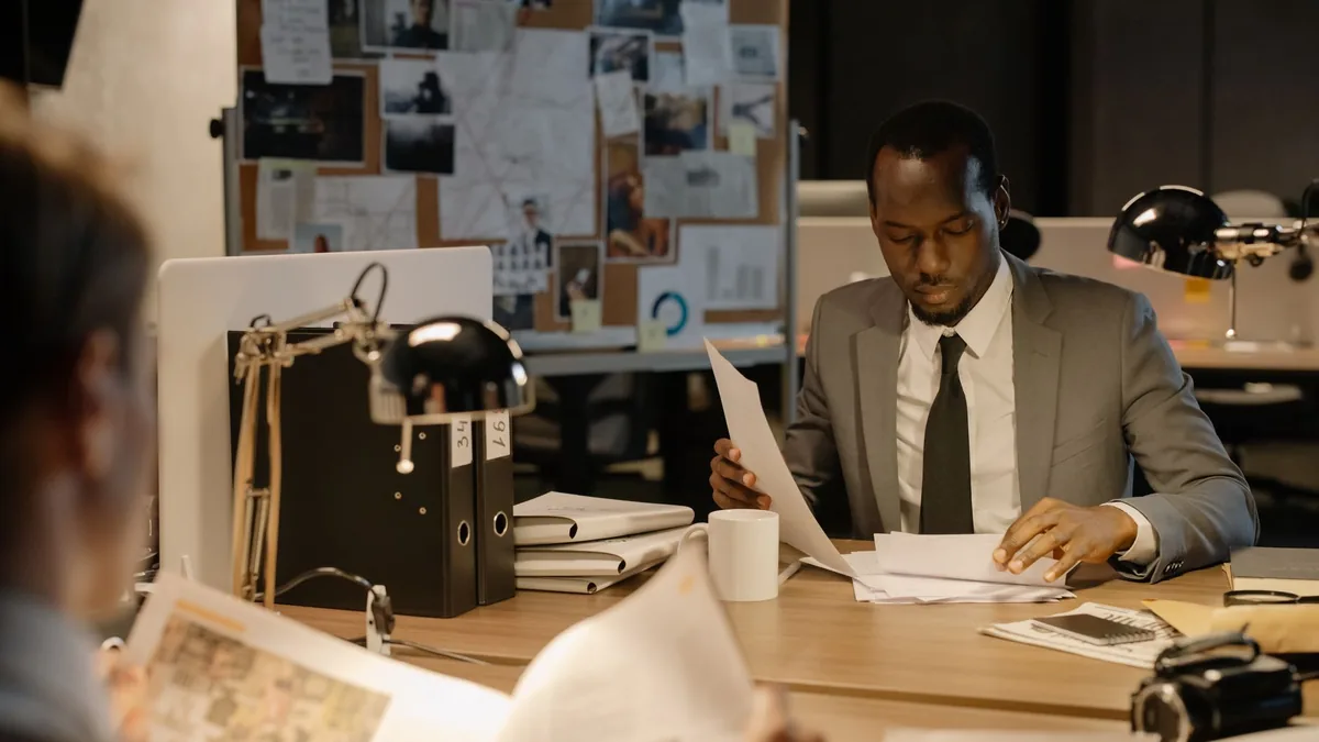 Detective in a suit examines papers at desk in a dimly l