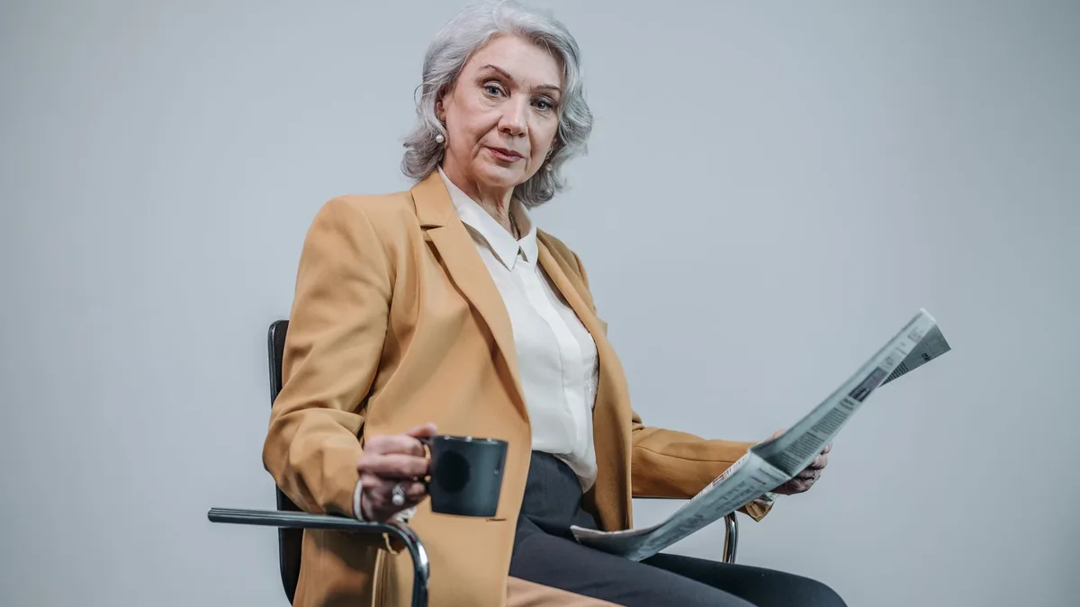 Elderly woman in brown blazer holding a newspaper and cu