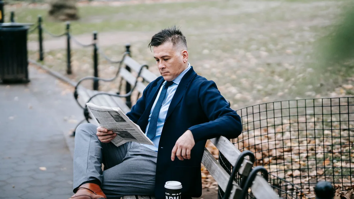 Man in formal attire reading a newspaper on a park bench