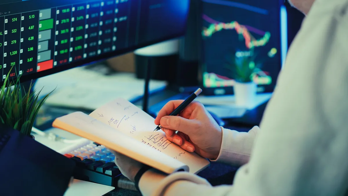 Person analyzing financial data on screens making notes