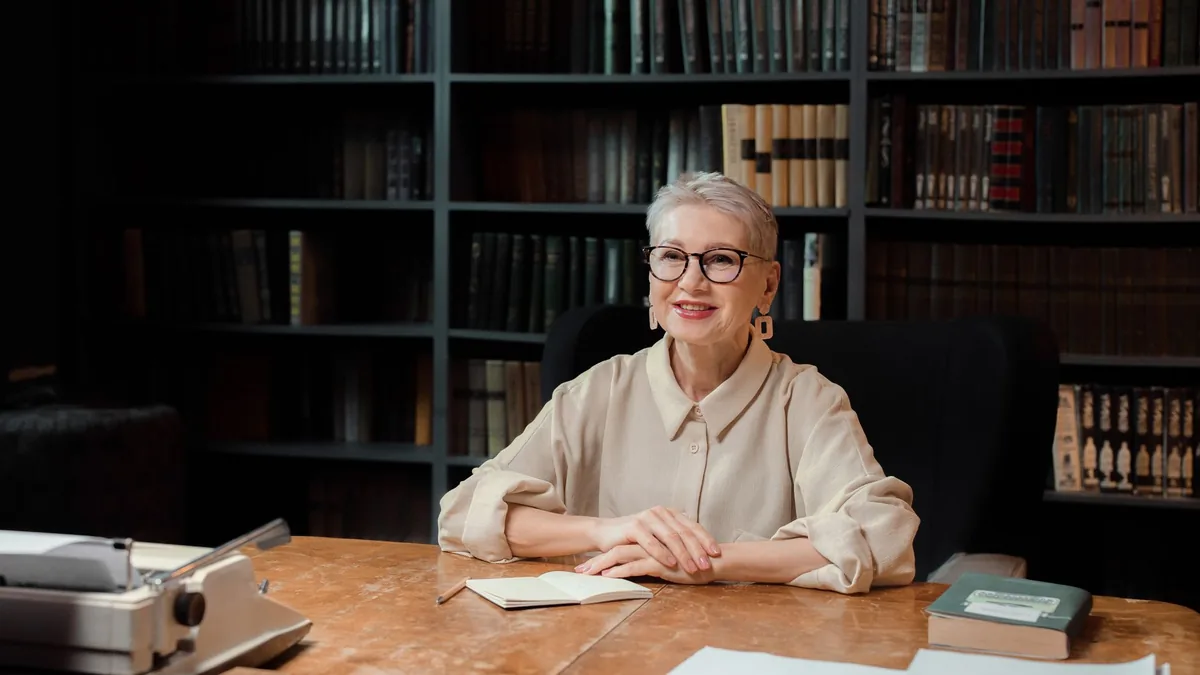 Senior woman sitting at desk in library smiling at camer