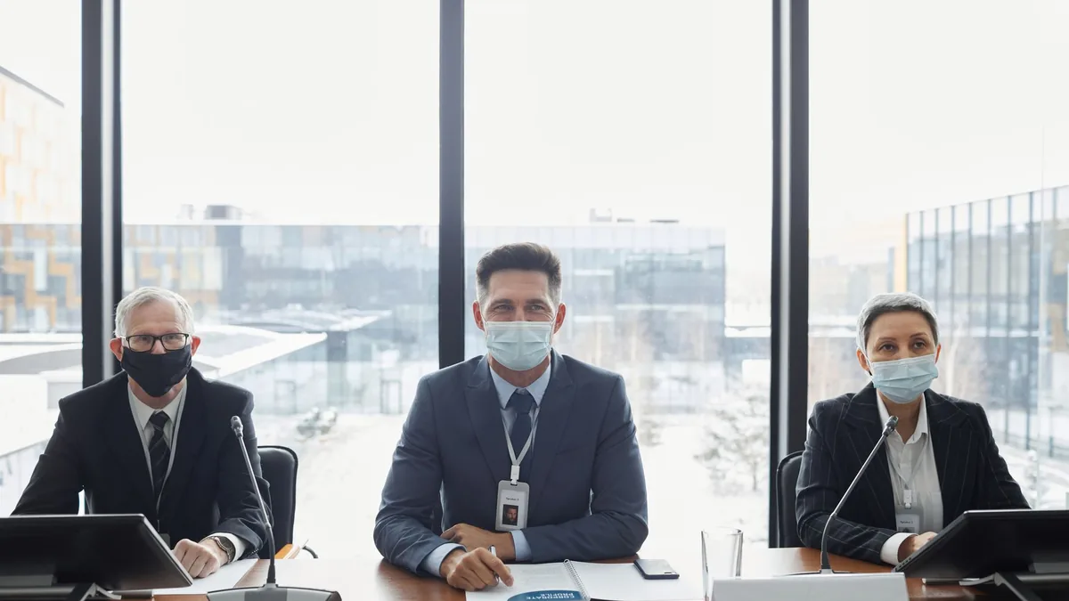 Three professionals in suits and masks sitting at a conf