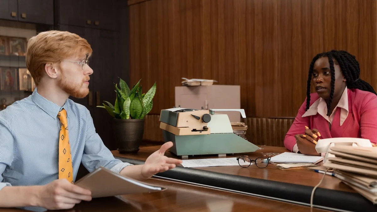 Two colleagues converse at a wooden office desk with vin