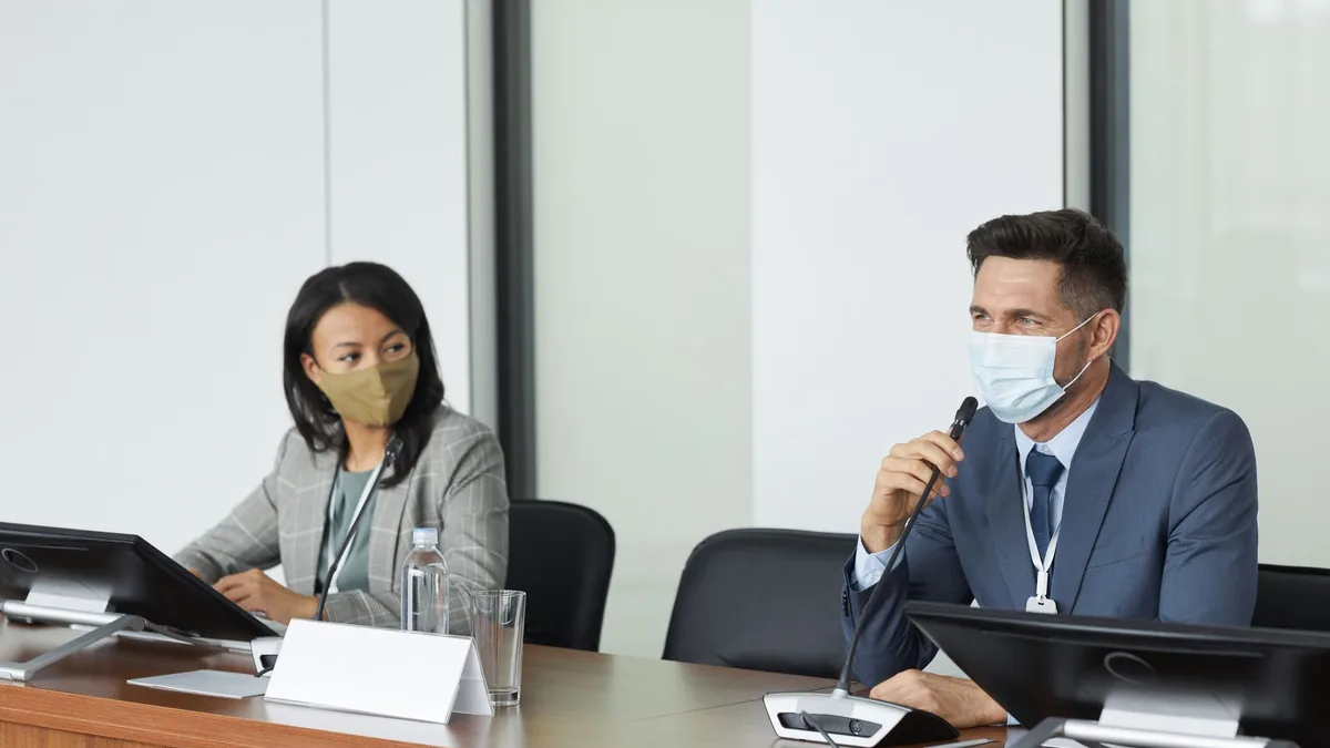 Two masked professionals in a conference room discussing